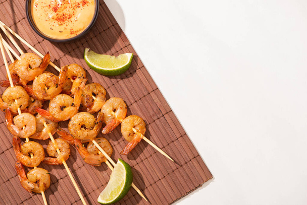 top view of prawns on skewers with lime and sauce on bamboo mat on white background - Foto, afbeelding