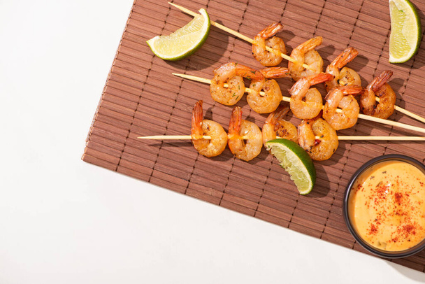 top view of prawns on skewers with lime and sauce on bamboo mat on white background - Zdjęcie, obraz
