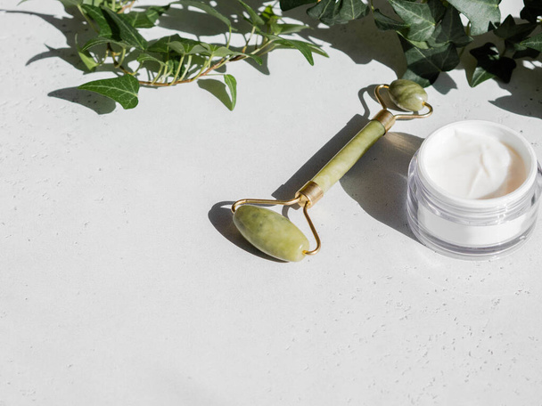 Yeşil Gua sha masaj yeşim taşı silindiri ve beyaz arka planda beyaz konteynırda yüz kremi. Boşluğu kopyala - Fotoğraf, Görsel