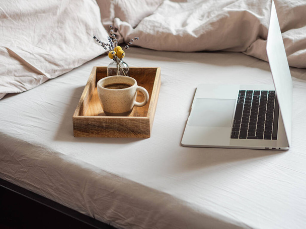 Laptop und Kaffee auf dem Bett im Schlafzimmer. Arbeit von zu Hause aus oder ein bequemer Zeitvertreib im Internet. Bleibt zu Hause, Quarantäne. Konzept Arbeit zu Hause. - Foto, Bild