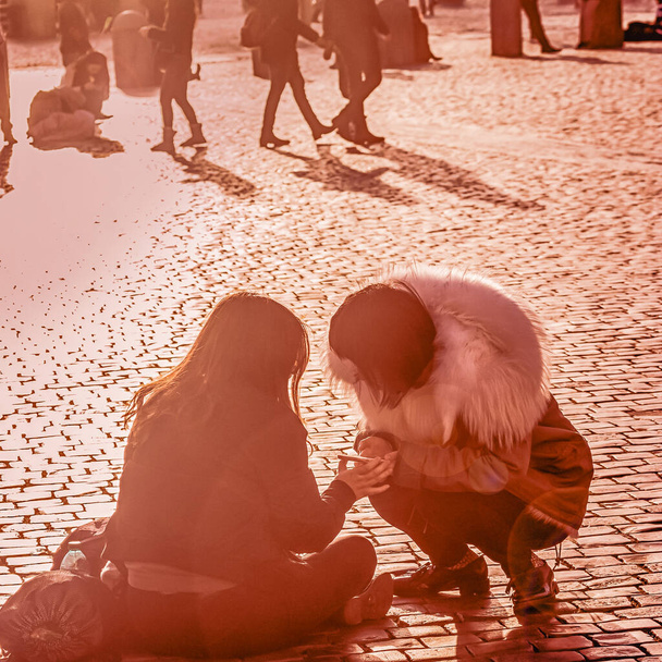Two girl friends sitting on the pavement and looking on one of their smartphones. Sunlight, coral color toned photo - Foto, afbeelding