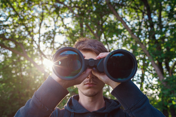 Explorer hiker young man in plaid shirt looking through binoculars on background of summer forest and sea bay outdoor, front view. Travel concept. - Foto, imagen