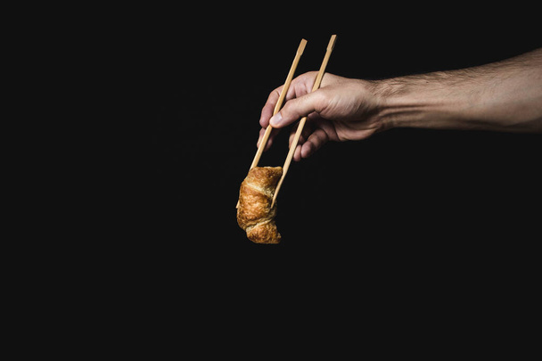 Hand holds croissant with chinese chopsticks on black background - Photo, image