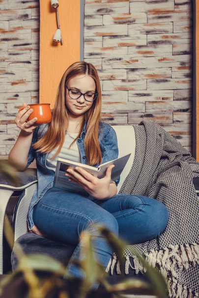 Happy teenage girl reading book and drinking tea in armchair in morning - Zdjęcie, obraz