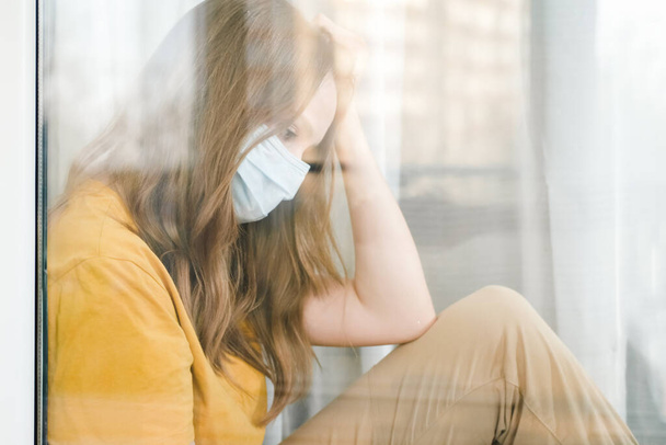 Sad girl in medical protective mask and yellow t-shirt sitting on the windowsill during a pandemic quarantine coronavirus Covid-19. Isolation at home. - 写真・画像