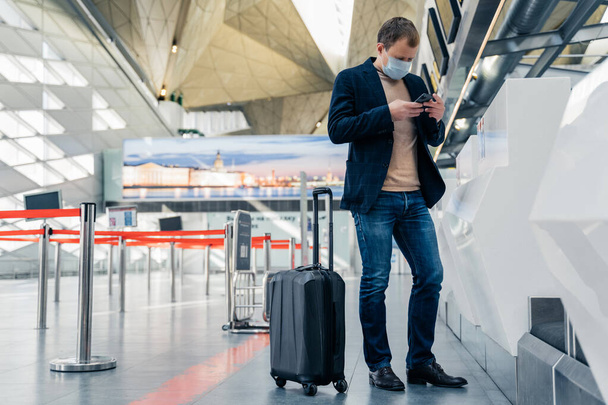 Täyspitkä laukaus mies matkustaja seisoo lähellä matkalaukku pyörillä lentokentällä lukee uutisia verkossa matkapuhelin kuluu suojaava lääketieteellinen naamio tietää sulkemisesta maiden rajojen takia virus
 - Valokuva, kuva
