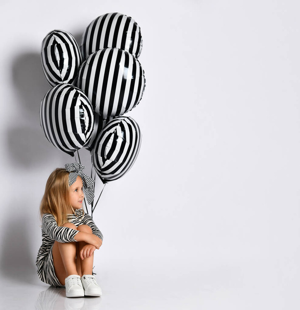 Schoolgirl in dress, headband and sneakers. Smiling, sitting on floor, posing with striped balloons isolated on white. Close up - Fotó, kép