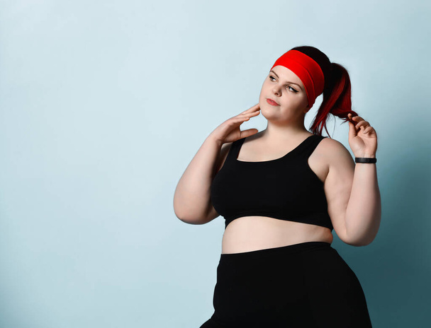 Concentrated plus-size girl in black sportswear stands looking aside thoughtfully, touching her face and holding ponytail in hand on blue background - Fotoğraf, Görsel