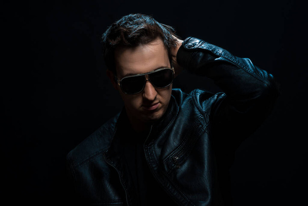 Young sexy stylish guy with glasses in a black leather jacket on a black background. studio shoot - Fotó, kép