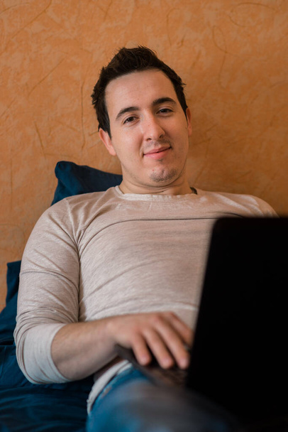 Een man die laptop gebruikt in een hotelkamer of slaapkamer. Hij ligt op bed en typt vanaf een laptop. Man in de ochtend met een modern apparaat, het lezen van e-mails en het verzenden van berichten. Afstandsonderwijs tijdens de Covid-19 pandemie. freelasner, ver weg  - Foto, afbeelding