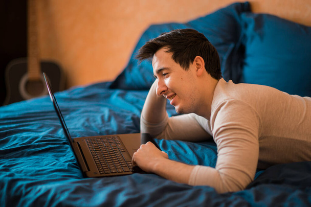 Man using laptop in a hotel room or home bedroom. The guy is lying on the bed and typing from a laptop. Man in the morning with a modern device, reading emails and sending messages. Distance learning during the Covid-19 pandemic. freelasner, distant  - Foto, immagini
