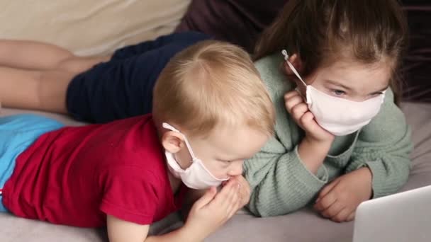 Two children are lying on a sofa at home in white medical masks and watching videos on a laptop. Concept of quarantine, self-isolation and epidemic. Video - Footage, Video