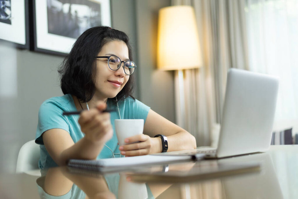 Beautiful Asian professional woman with blue shirt and eyeglasses in confident expression while work from home. Attractive young girl working on laptop computer during quarantine and isolated. Prevention Pandemic virus. Covid 19, Coronavirus, Healthy - Фото, зображення
