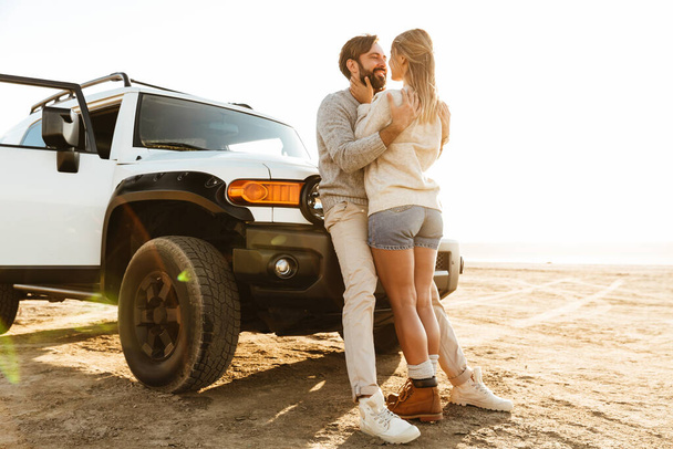 Feliz joven pareja enamorada abrazándose mientras se apoya en un coche en la playa soleada
 - Foto, Imagen
