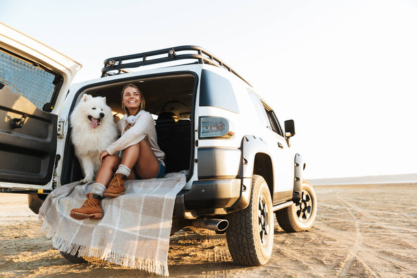 Cheerful lovely girl playing with her dog while sitting in a car at the beach - Foto, Imagem