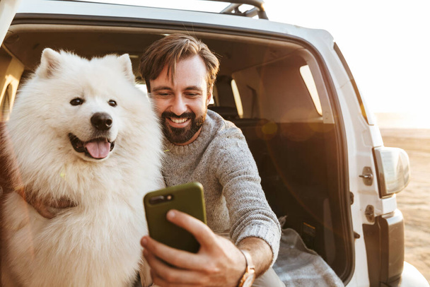 Przystojny młody brodaty mężczyzna siedzący z tyłu samochodu, bawiący się psem na plaży, robiący selfie - Zdjęcie, obraz