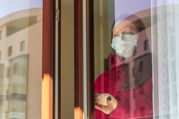 Κορίτσι ή νεαρή γυναίκα με ιατρική μάσκα που βρίσκεται σε καραντίνα στο σπίτι κατά τη διάρκεια της πανδημίας του κορωναϊού. Αυτοαπομόνωση, θέα μέσα από γυαλί, περπάτημα στο μπαλκόνι ενός διαμερίσματος στην πόλη - Φωτογραφία, εικόνα