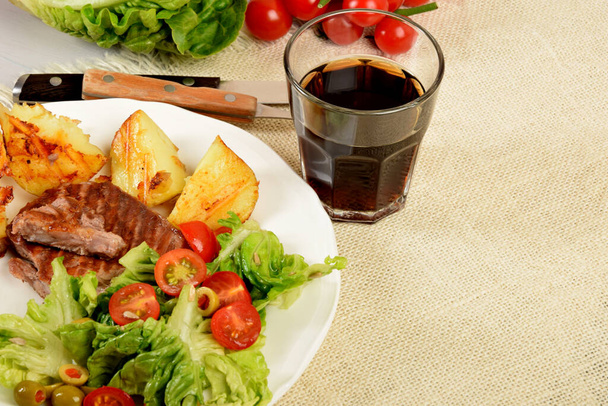 pieces of grilled steak next to potatoes grilled next to lettuce with tomatoes and olives and a drink in a glass - Foto, Imagem