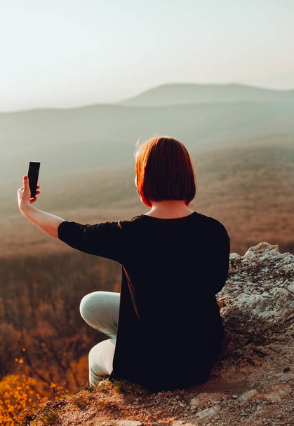 Młoda atrakcyjna kobieta trzymająca smartfona i robiąca selfie na skale, nad doliną z doskonałym widokiem na zachód słońca. Dziewczyna na szczycie robi sobie zdjęcie z przygody - strzał z tyłu. - Zdjęcie, obraz