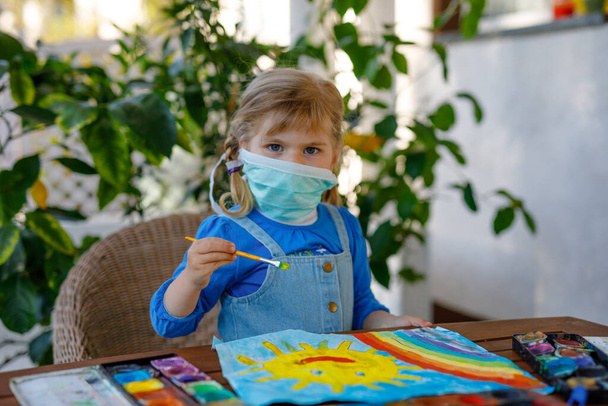 little toddler girl in medical mask painting rainbow with water colors during pandemic coronavirus quarantine disease. Children painting rainbows around the world with the words Lets all be well. - Photo, Image