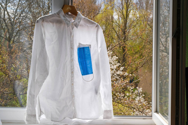 Pure white shirt and blue face mask. Shirt hanging on the background of the open window. Self-isolation. - Fotoğraf, Görsel