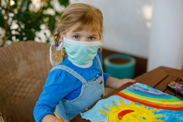 Mała dziewczynka w masce medycznej maluje tęczę kolorami wody podczas pandemii choroby kwarantanny koronawirusowej. Dzieci malujące tęcze na całym świecie słowami Niech wszystko będzie dobrze. - Zdjęcie, obraz