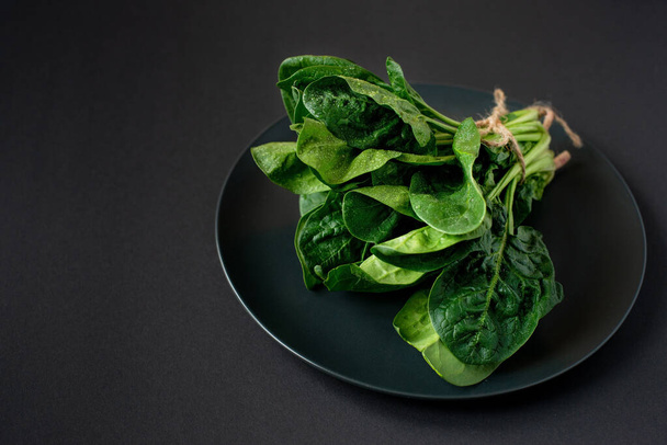 Clean food concept. Bunch of leaves of fresh organic spinach greens in a plate on a black background. Healthy detox spring-summer diet. Vegan Raw Food. - Foto, Imagen