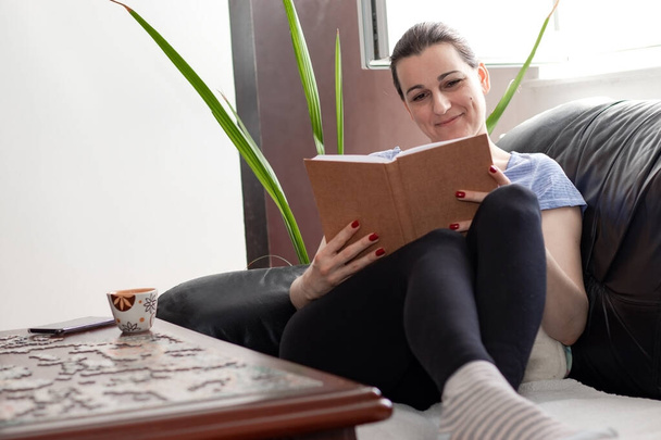 Egy fiatal nő ül a lábával a kényelmes kanapén és könyvet olvas. Maradj otthon koncepció idején világjárvány COVID 19 vírus. - Fotó, kép