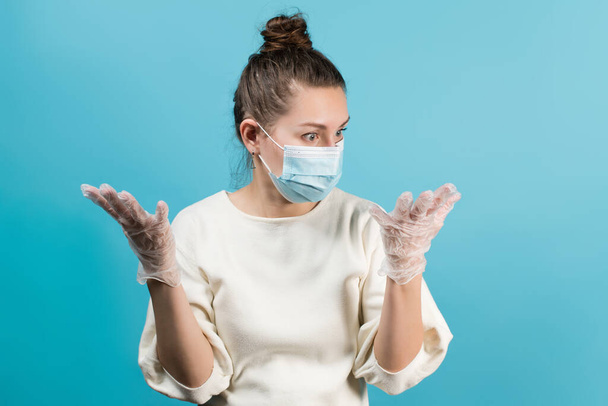 A woman in a medical mask looks at her hands with medical gloves in surprise and shock. Isolated on a blue background - Φωτογραφία, εικόνα