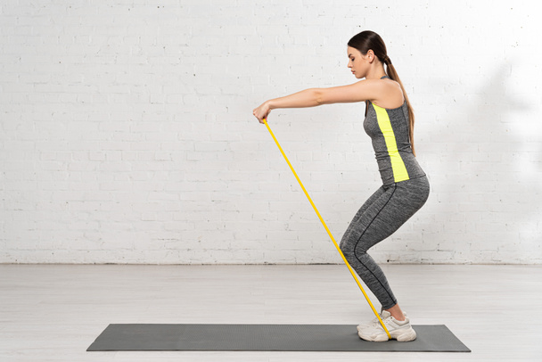 side view of woman in sportswear working out with resistance band on fitness mat near white brick wall  - Photo, Image