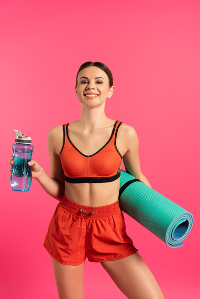 happy sportswoman holding sports bottle with water and fitness mat isolated on pink  - Фото, изображение
