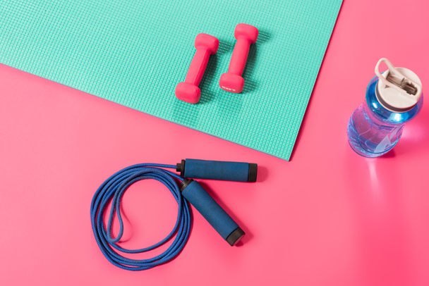 top view of sports bottle with water near skipping rope and dumbbells on fitness mat on pink  - Foto, afbeelding