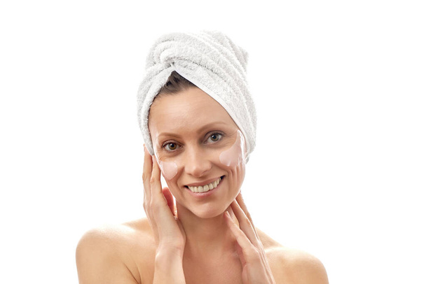 A beautiful woman with a white towel on her head does facial rejuvenation with eye patches and is smiling at the camera. White isolated background - Fotó, kép