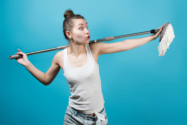 A young girl amusingly holds a mop around her neck like a rockstar microphone stand at a concert. Fun cleaning concept. Isolated on blue - Foto, Imagem