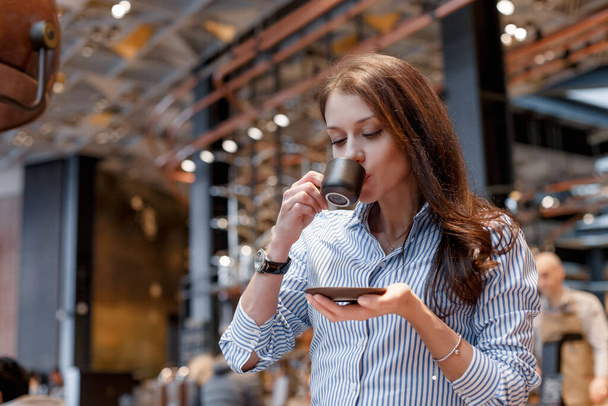 Young beautiful girl in a beautiful modern cafe drinks delicious coffee. Girl in a fashionable blue shirt - Foto, Imagen