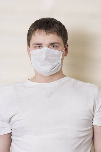 A man in a white T-shirt and a medical mask stands on a light background - Foto, Imagem