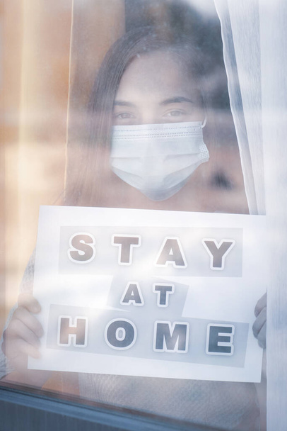 Concept of coronavirus quarantine. Child wearing medical protective face mask during flu virus, looking out of window. COVID-19. Teen girl holds board sign with text Stay at home. View from street. - Foto, afbeelding