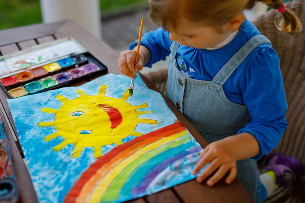 Маленькая девочка рисовала радугу и солнце акварелью во время пандемического коронавирусного карантина. Дети рисуют радуги по всему миру со словами "Давайте все будем здоровы". Счастливый ребенок
 - Фото, изображение