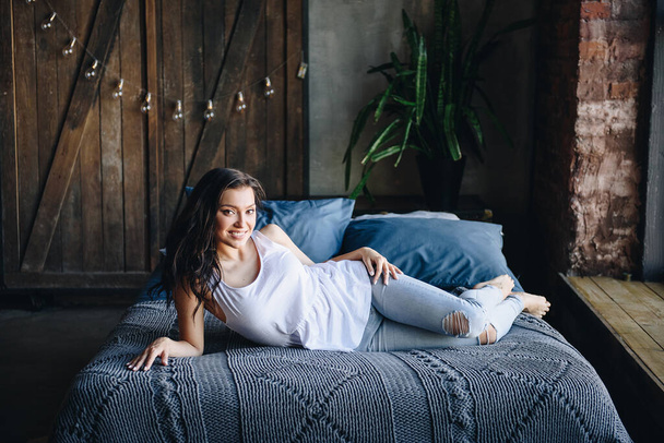 Beautiful brunette girl. Eastern appearance. In jeans and a white T-shirt. Sitting at home on the couch, lying on the bed. Home furnishings. Portrait. Slim and fit figure. Gray sofa. Interior loft. - 写真・画像