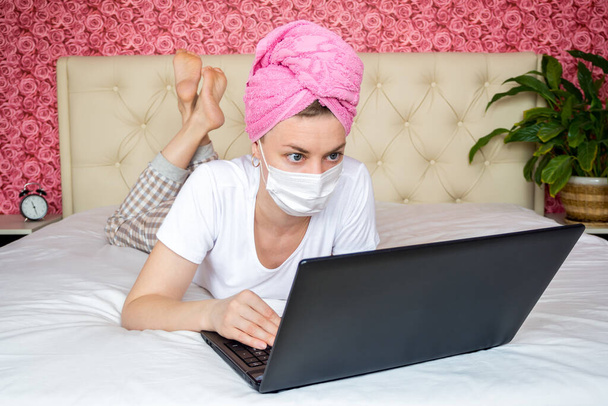 Remote work from home quarantine. Young Caucasian girl blogger working on a laptop on a bed in a mask. remote communication, online shopping, home orders during the pandemic - Fotografie, Obrázek