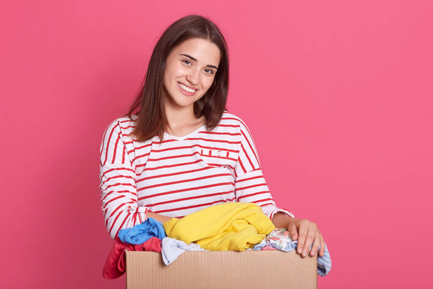 Horizontal shot of smiling brunette woman dresses striped casual shirt, looking directly ar camera with happy facial expression, carrying carton box with donate clothing, lady doing charity work. - Φωτογραφία, εικόνα