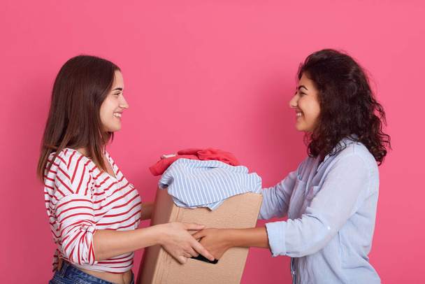 Indoor shot of two attractive women posing isolated over rosy background, smiling lady handing big carton box with clothes, young female gives charity clothing for poor people. Donation concept. - Fotó, kép