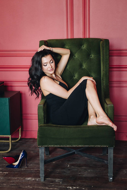 Beautiful girl with oriental appearance. Black fitting dress on the straps. Long dark hair. Red wall. Barefoot. Green chair. Yellow flowers. Sexy and bright woman. Portrait. - Foto, afbeelding