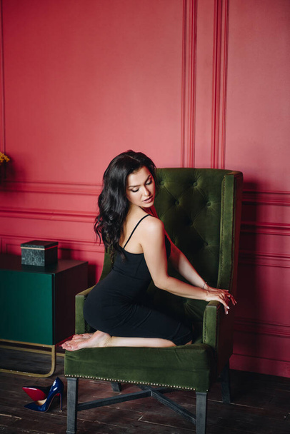 Beautiful girl with oriental appearance. Black fitting dress on the straps. Long dark hair. Red wall. Barefoot. Green chair. Yellow flowers. Sexy and bright woman. Portrait. - Фото, зображення
