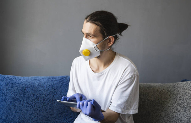 Man in white shirt, medical mask and rubber gloves sits at home and works with tablet on a sofa during quarantine. Man, designer, artist, architect, businessman at remote work in a pandemic covid - Foto, afbeelding
