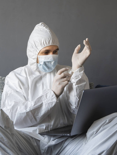 Man in protective suit, medical mask and rubber gloves sits at home and works with laptop at the table during quarantine. Designer, artist, architect, businessman at remote work in a pandemic covid. - Foto, immagini