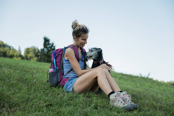 Cheerful young woman sitting in green grass cuddling her black dog as the take a rest during  hike. - Zdjęcie, obraz