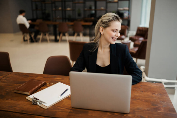 A business lady in a classic suit sits at a wooden table with a laptop and looks out the office window with a smile - Foto, imagen