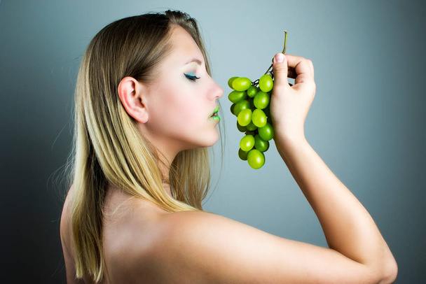 Piękna dziewczyna trzymająca winogrona w rękach. Dziewczyna wyglądająca jak modelka będzie jadła zielone winogrona. Zielone usta dziewczynek harmonizują z zielonymi winogronami. - Zdjęcie, obraz