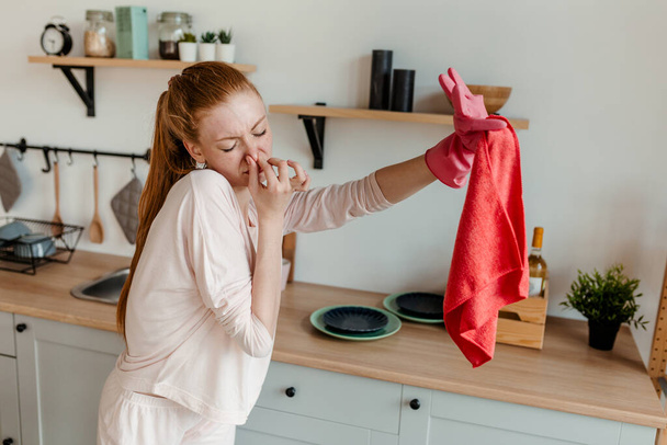 Red-haired young girl clamps her nose against the strong smell of a rag while cleaning the kitchen - Foto, Bild